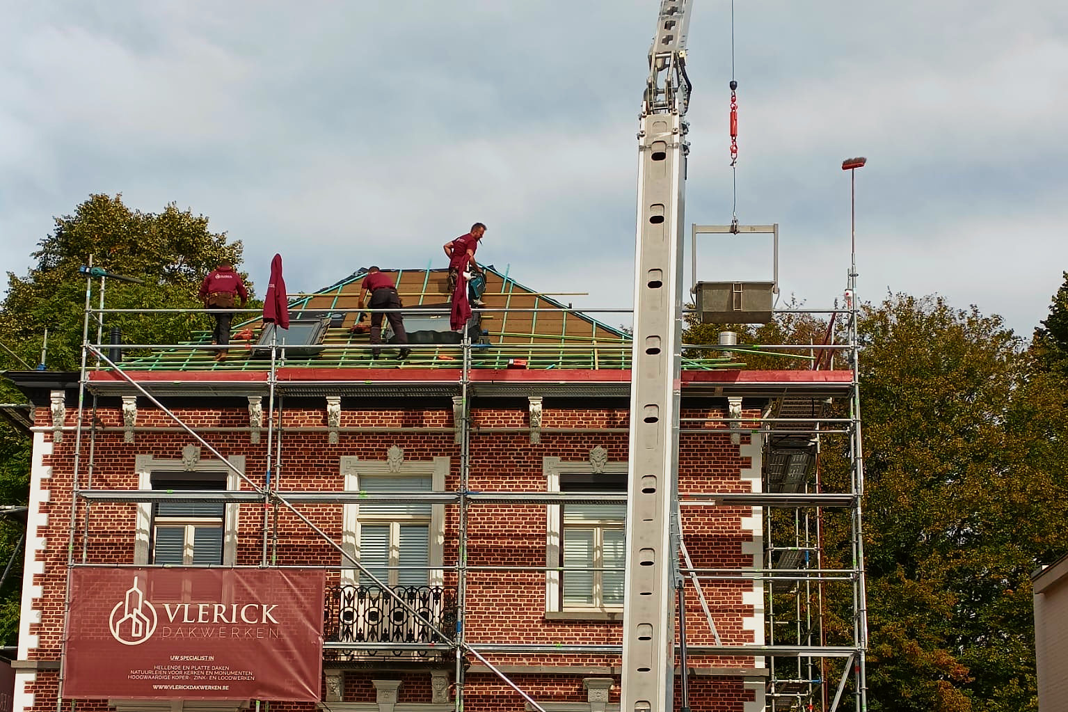 Het bakstenen herenhuis in de stelling, met een kraan en vakmannen op het dak.