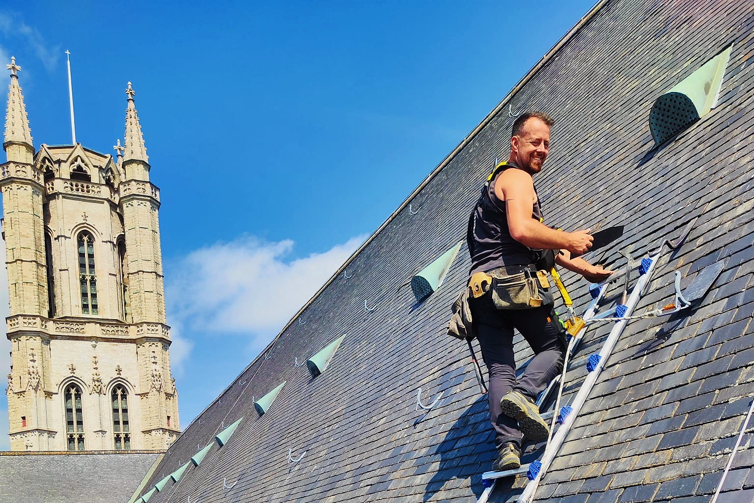Een vakman van Vlerick Dakwerken op een dakladder op het steile leiendak van de Sint-Baafskathedraal, met de toren op de achtergrond.