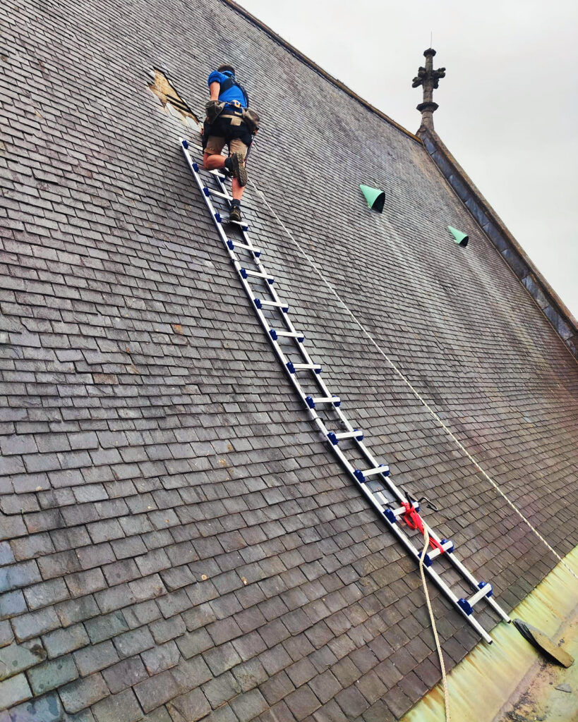 Een vakman aan het werk bovenaan een dakladder op het grote leiendak, gezekerd met een veiligheidslijn.
