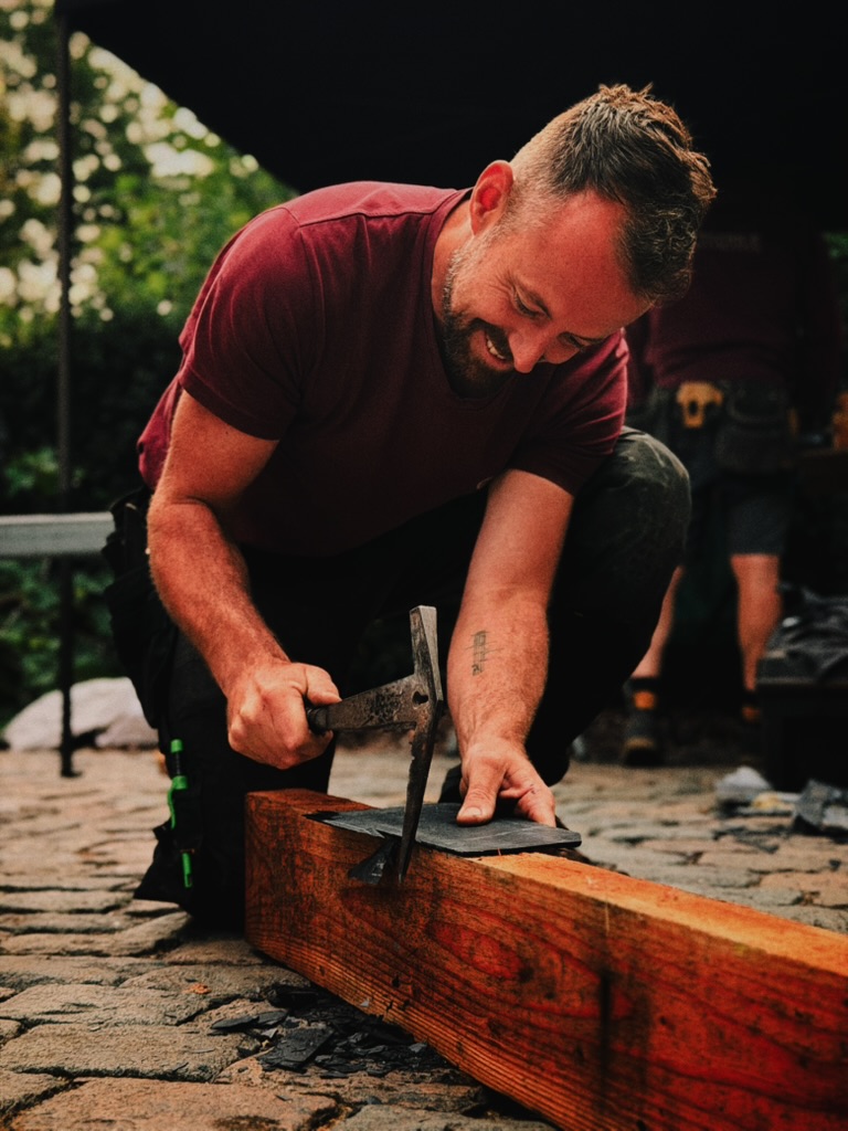 Een vakman van Vlerick Dakwerken kapt met de leihamer een natuurlei op maat op een houten balk.